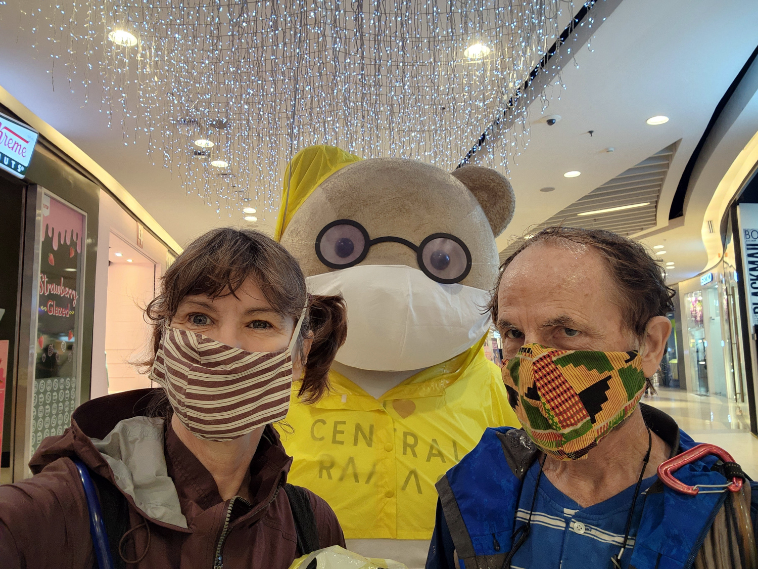 two travel bloggers posing for a photo with a giant teddy bear while getting out of the rain. At a mall in Bangkok