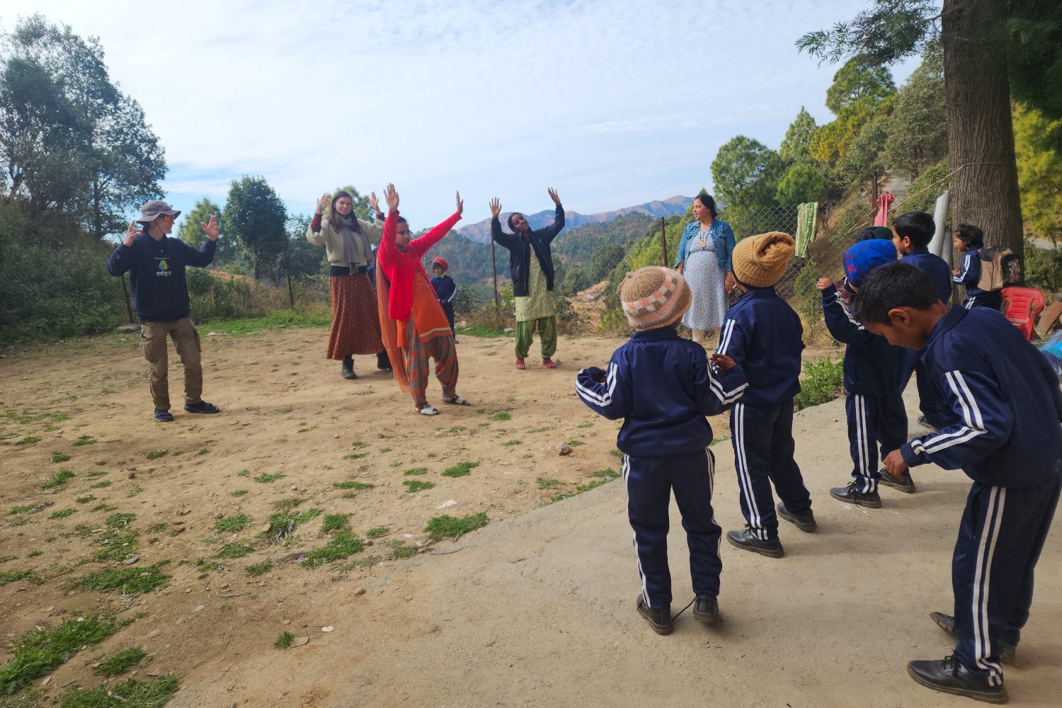 Children in blue uniforms participate in an outdoor activity with adults in a sunny Himalayan village, raising their hands in excitement or instruction. The scene features a mix of dirt ground and a concrete slab, with lush green trees and distant mountains under a partly cloudy sky.